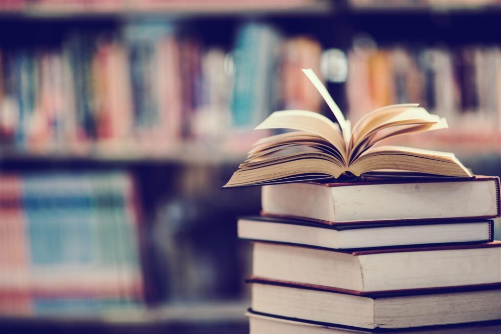 Livro aberto sobre uma pilha de livros em frente a uma estante